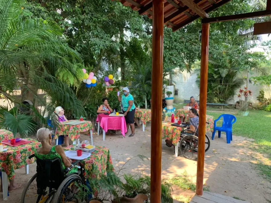Residentes reunidos em mesas ao ar livre no jardim, durante uma festa com balões, acompanhados pela equipe